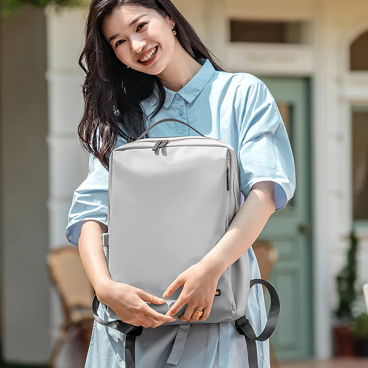 明るい日差しの下、白い柱とミント色のドアがある家の前で、青いシャツワンピースの女性が軽量で上品なビジネスリュックを手に持ち微笑んでいます。